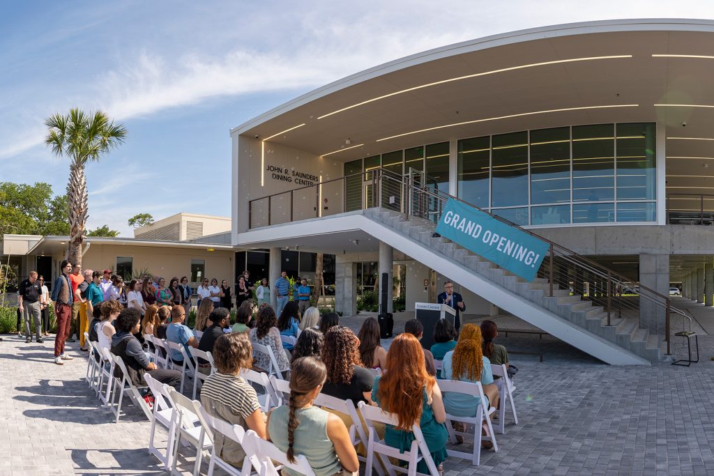 A group of spectators stands outside of a modern two-story building to listen to a man in formal attire addressing the crowd from a podium