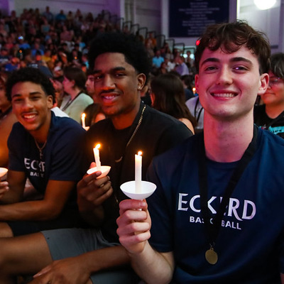 Three students wearing shirts that read ECKERD sit side by side holding candles and smiling in an auditorium with dimmed lights