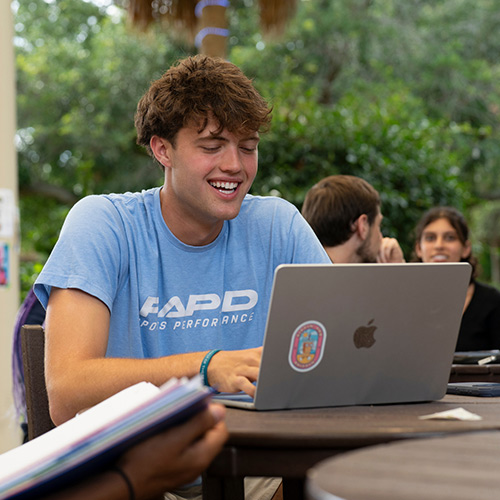 Student writing on a laptop while seated outside with other students