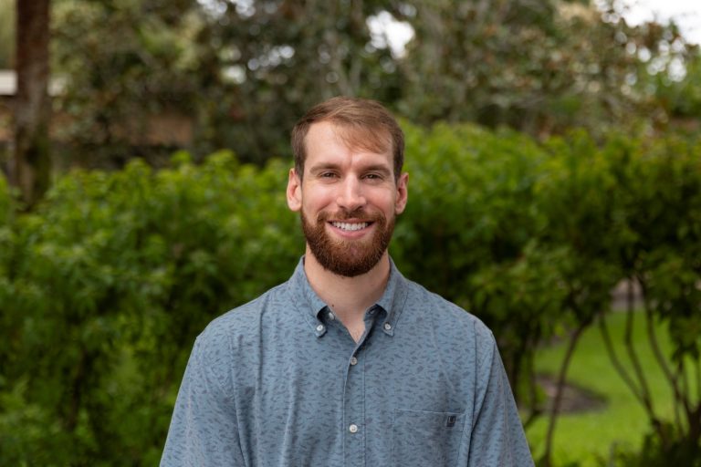 A smiling man standing in front of a background of green healthy shrubbery.