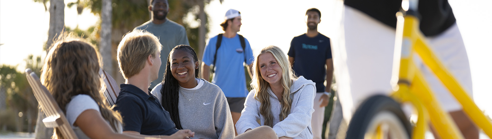 Four students sit in a group outside on the beach with morning sun hitting them and a yellow bike in the foreground
