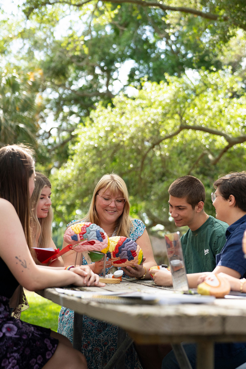 Professor shows students a model of a brain while seated around a picnic table outside under trees