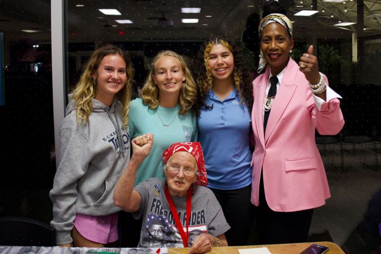Four women stand behind one elderly woman in a red bandana who is seated and raising her fist.